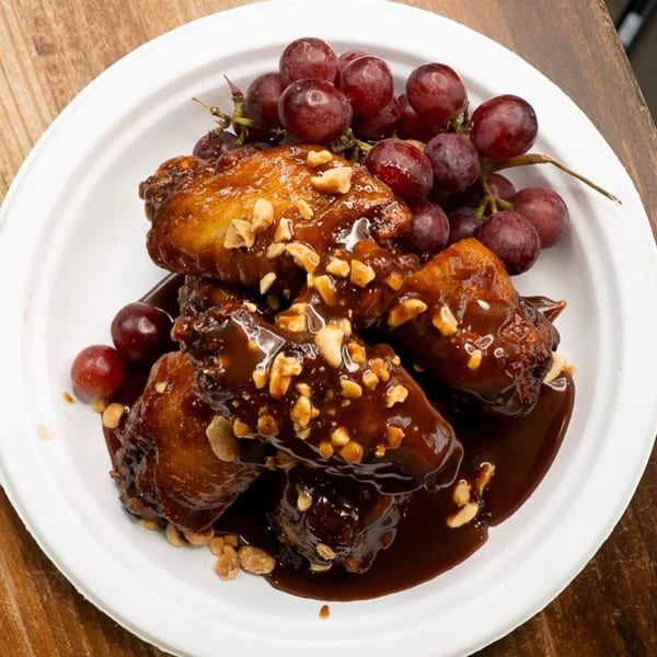 King's Chicken Wings, glazed with sauce and peanuts, served with red grapes on a white plate.