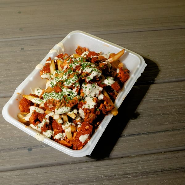 King's Chicken Wings' fries topped with red chicken pieces, white sauce, and green herbs in a styrofoam container.